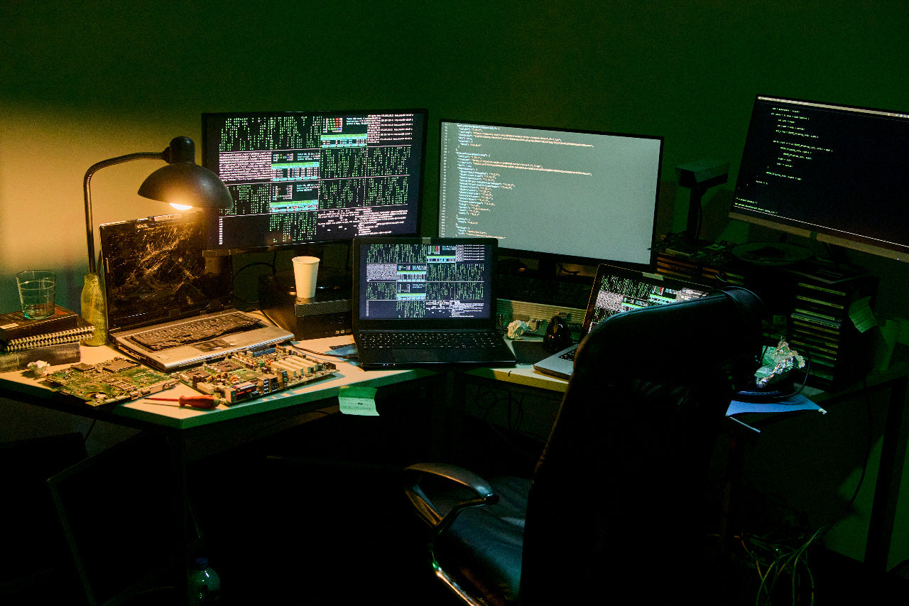 Dim desk setup with multiple monitors showing code, a lamp, and disassembled computer parts