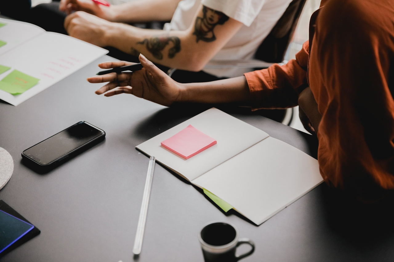 Close up of a tattooed arm reaching across a desk with notebooks, a phone and a coffee cup
