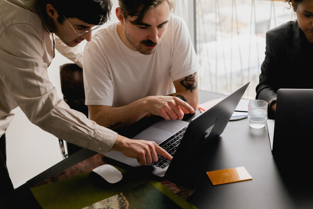 Two coworkers lean over a laptop at a desk while a third looks on in the background