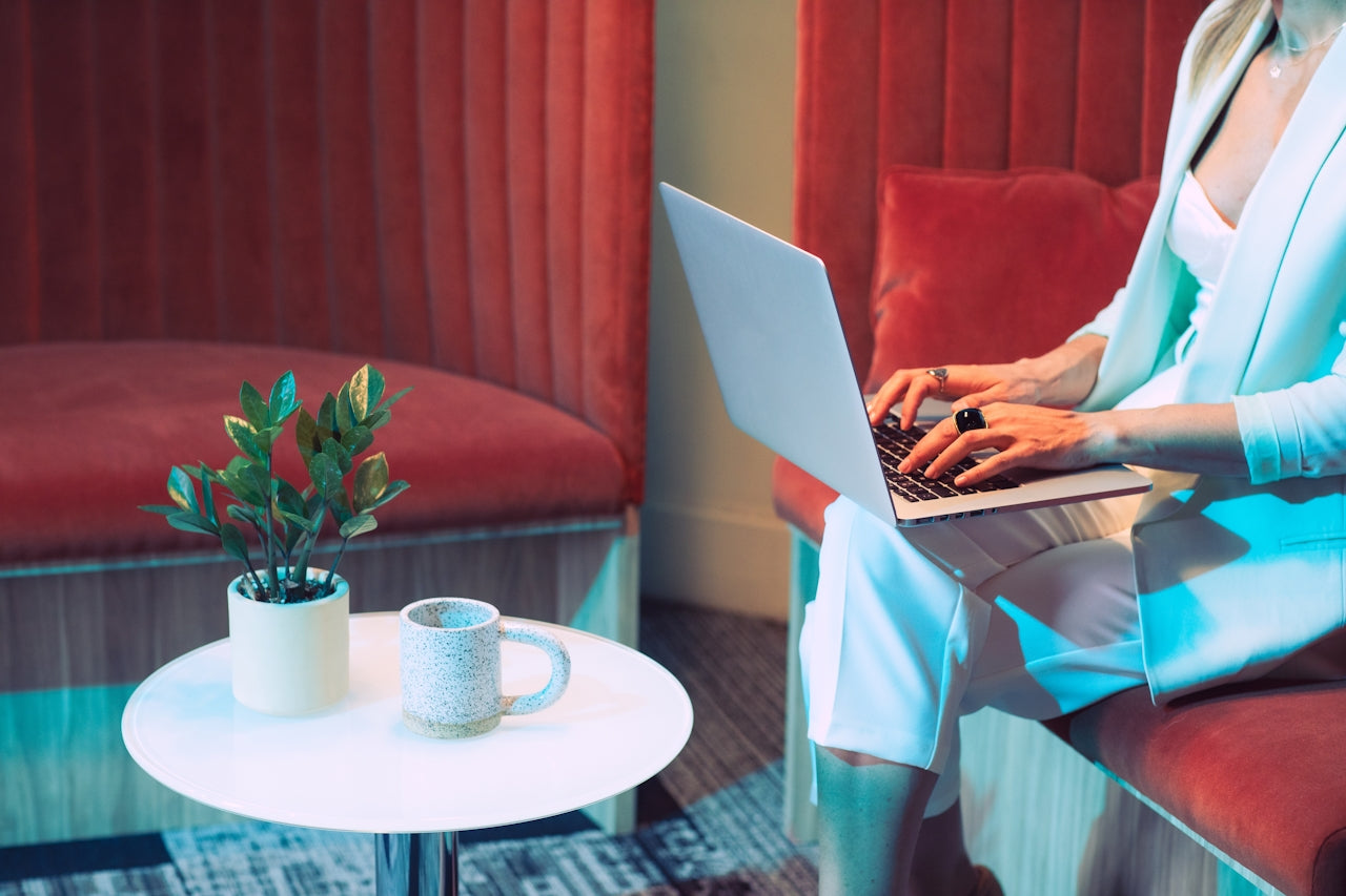 Woman works on a laptop in a lounge booth with red velvet seating and a small plant