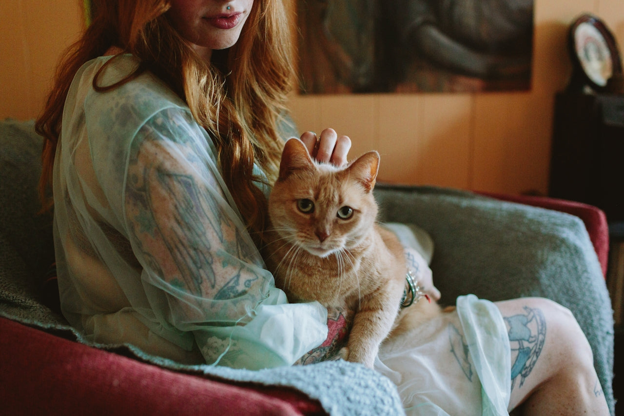 Woman with tattoos sits on a couch petting an orange tabby cat on her lap
